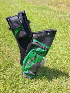 A golf bag standing upright on green grass under sunlight.