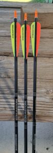 Three black carbon arrows with orange and yellow fletching on a wooden surface.