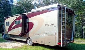 A beige and maroon Endura RV parked on grass with a ladder at the back.