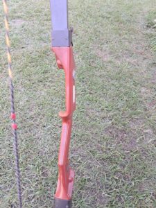 A wooden recurve bow with a red grip, lying on grass beside a fishing rod.