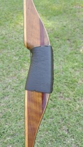 Close-up of a wooden bow grip with a black textured handle on grass.