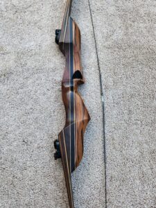 A wooden recurve bow with a stabilizer on a concrete surface.